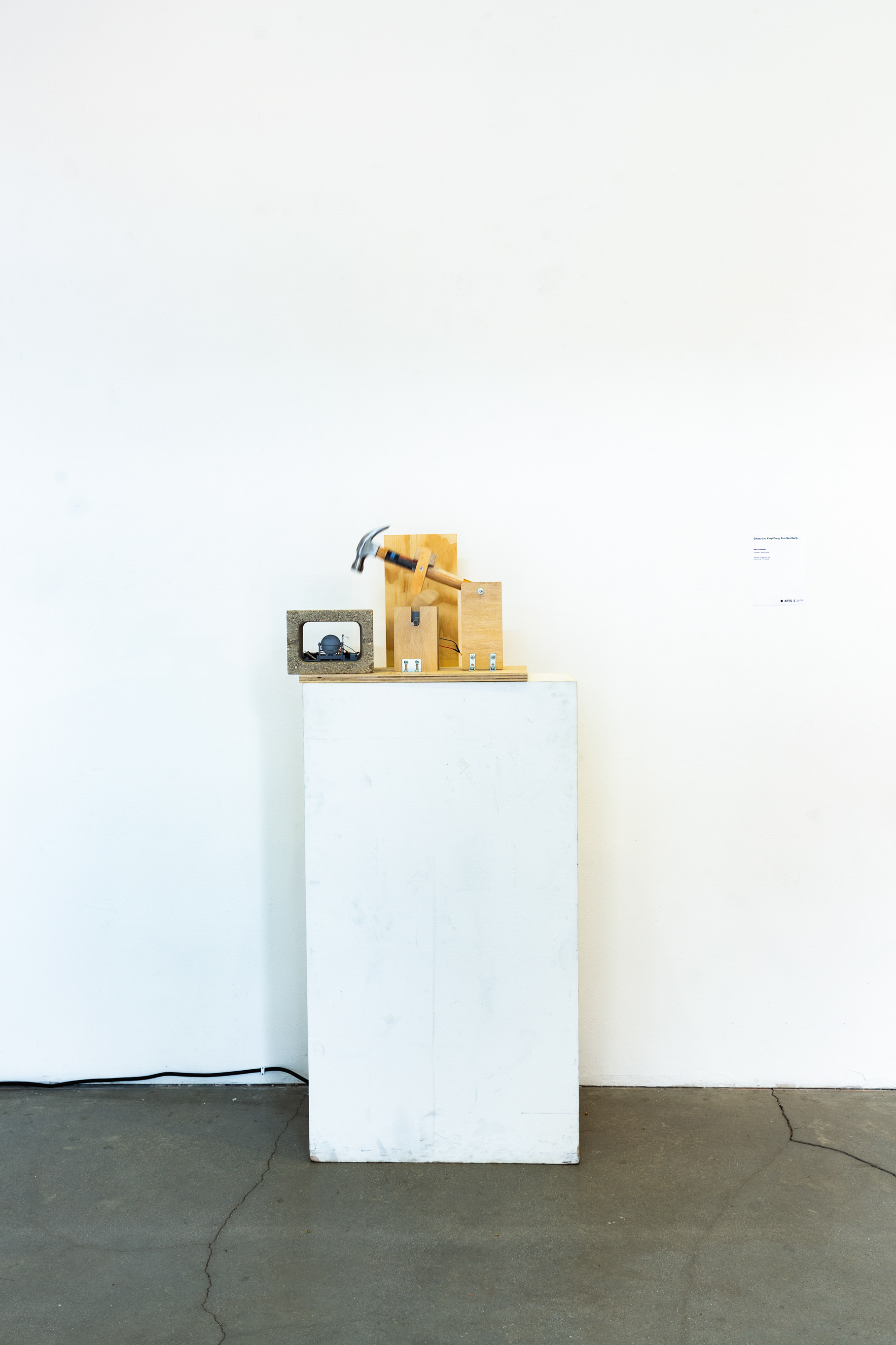 A sculpture consisting of a hammer assembeled by wood with electronics, where the hammer strikes a cinderblock, on a white podium against a white wall.